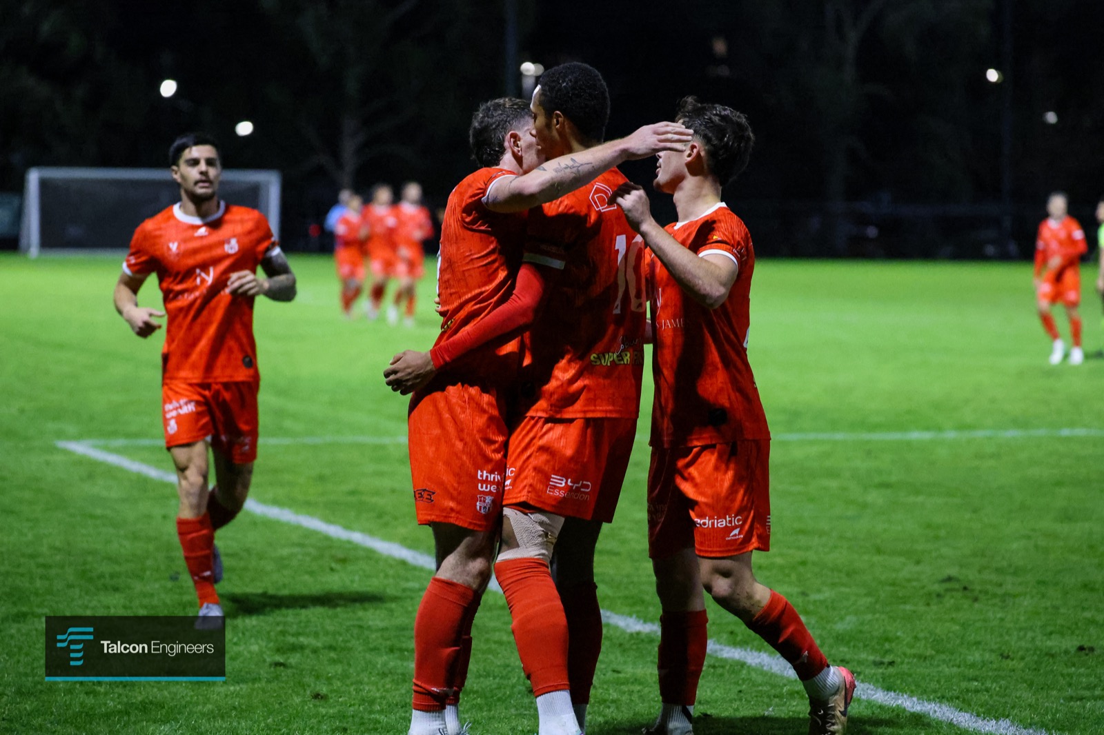 Community football club players celebrating a goal under lights, sponsor logos on the kit